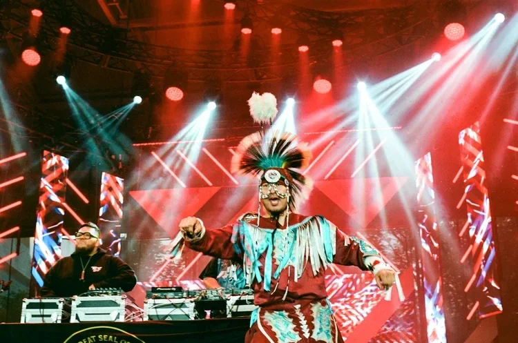Performer in traditional regalia on a concert stage lit with red and teal lighting