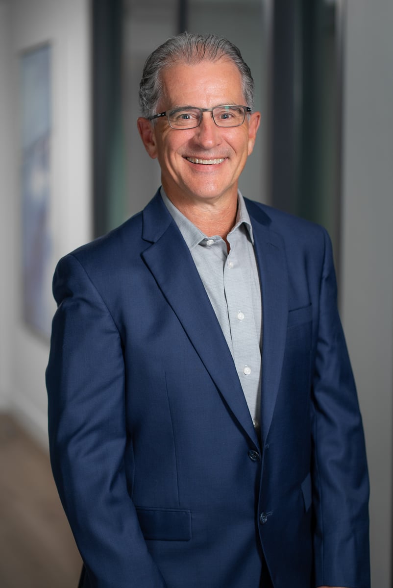 Corporate headshot of a smiling executive in a navy blazer against a dark backdrop